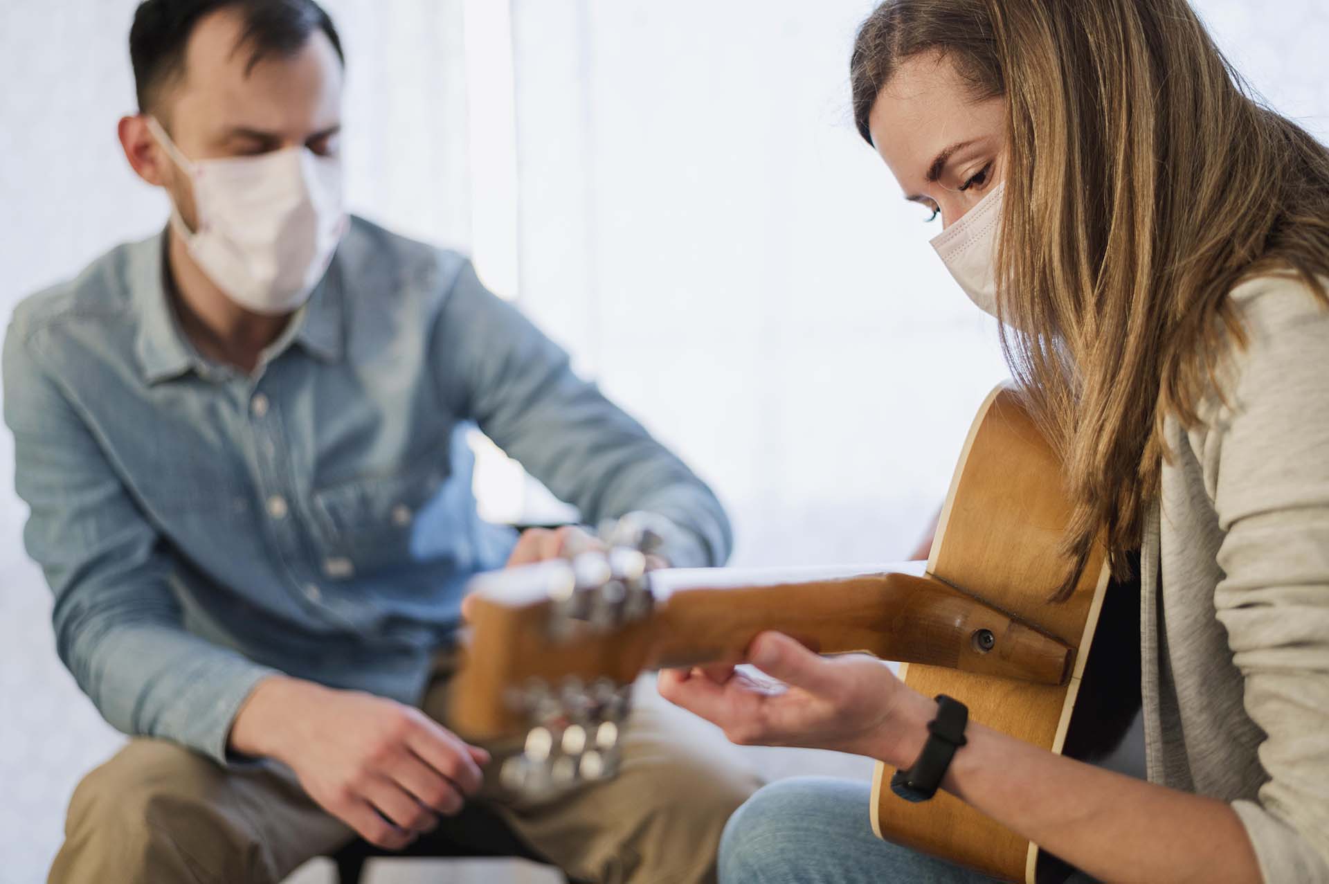 Man with a face mask relaxing and instructing woman who also has a facemask on how to play the guitar as therapy for her addiction recovery